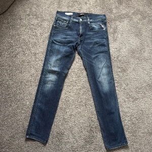 Replay Anbass Hyperflex  - Snygga Replay Anbass Hyperflex jeans i mörkblå tvätt med slitningar och kontrastsömmar. Slim fit-modell med klassiska fem fickor, dragkedja och knappstängning. Stretchigt material som ger skön passform och rörelsefrihet. Perfekt för dig som gillar stilrena jeans med edge.