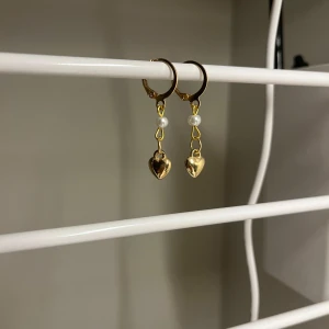 Guldiga örhängen med hjärtan och pärlor - Supersöta örhängen i guldton med hängande hjärtan och små vita pärlor. Perfekta för att ge din look lite extra charm och romantisk vibe. Passar dig som gillar detaljer och vill sticka ut med något gulligt och trendigt. I rostfritt stål och handgjort