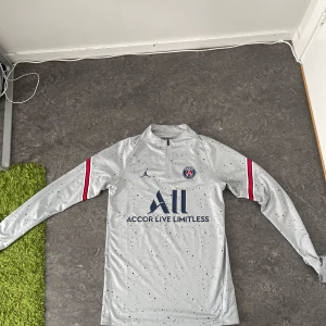 PSG långärmad träningströja Jordan - Snygg PSG träningströja från Jordan i ljusgrått med mörkblå prickigt mönster. Långärmad modell med halv dragkedja, PSG-logga på bröstet och Accor Live Limitless-tryck. Röd och blå rand på ärmarna, tillverkad i mjukt och stretchigt funktionsmaterial.