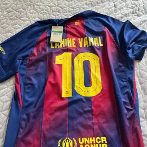 FC Barcelona Lamine Yamal tröja Nike - Säljer en officiell FC Barcelona matchtröja med Lamine Yamal och nummer 10 på ryggen. Tröjan är kortärmad, har klassiska blå och röda vertikala ränder, gula detaljer och klubbmärke på bröstet. Materialet är lätt och andas, perfekt för fotboll eller häng med vänner.