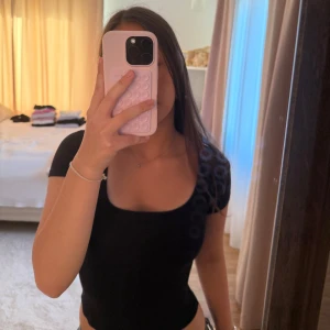 Svart kort tight kortärmad topp - Säljer en svart, tight topp med korta ärmar och djup rundad halsringning. Enkel och stilren design som sitter snyggt och framhäver figuren. Perfekt att matcha med jeans eller kjol för en clean look.