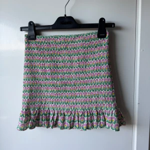 Blommig kortkjol med volang från Zara - Supersöt kortkjol från Zara med blommigt mönster i rosa, grönt, vitt och gult. Kjolen har smockad struktur och en volangkant längst ner som ger en extra girly vibe. Perfekt för dig som gillar färg och mönster! Elastisk midja för skön passform. Kan även användas som topp.