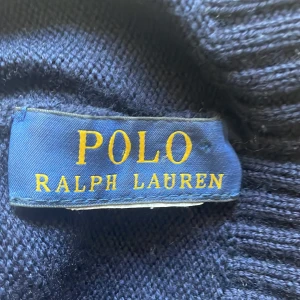 Mörkblå mössa Polo Ralph Lauren - Snygg mörkblå stickad mössa från Polo Ralph Lauren med klassisk röd broderad logga framtill. Mössan har ribbad kant och är tillverkad i mjukt material som håller dig varm och stilren under kyliga dagar.Perfekt för vintern och hösten. Passar dom flesta som inte har stora huvuden 
