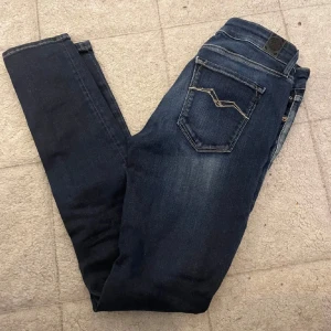 Replay Anbass s jeans blå - Säljer ett par Replay Anbass jeans i mörkblå tvätt med snygga slitningar och klassisk femficksdesign. Modellen är smal passform och låg midja. Jeansen har detaljerade sömmar på bakfickan och är tillverkade i stretchig bomull för extra komfort.