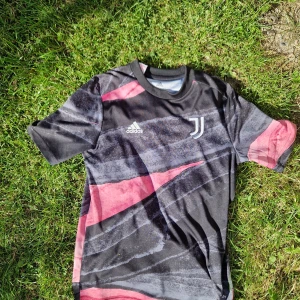 Juventus fotbollströja Adidas 152 - Juventus träningströja från Adidas i Aeroready-material. Tröjan har svart bas med rosa och gråa grafiska mönster, korta ärmar och rund hals. Juventus-logga och Adidas-märke på bröstet. Perfekt för träning eller match!