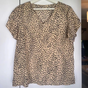 Leopardmönstrad beige blus - Snygg beige blus med svart leopardmönster och v-ringad hals. Blusen har korta ärmar och en loose passform som ger en chill vibe. Perfekt för dig som vill sticka ut med ett djuriskt print. Materialet känns lätt och luftigt mot huden.