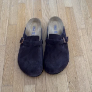 Birkenstock Boston  - Säljer ett par Birkenstock Boston sandaler i mörkbrun mocka med rund tå och justerbart spänne i metall. Klassisk slip-on modell med mjuk innersula och platt sula, perfekt för chill dagar. Materialet är mocka och innersulan är i skinn. Storlek 43. (Passar en 44 ingen låda)