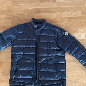 Mörkblå dunjacka från Moncler - Snygg mörkblå dunjacka från Moncler med quiltad design och hög krage. Jackan har dragkedja framtill, två fickor med dragkedja och ikonisk Moncler-logga på ärmen. Fylld med vitt gåsdun för extra värme och lätt känsla. Perfekt för kalla dagar och stilrena outfits.