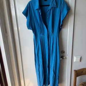 Blå långklänning från Gina Tricot - Snygg blå långklänning från Gina Tricot i storlek S. Klänningen har krage, knappar framtill och korta ärmar. Tillverkad i mjukt material med 55% lyocell och 45% viskos, vilket gör den skön och luftig. Perfekt för dig som gillar enkel och stilren design.