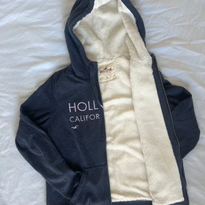 Mörkblå hoodie från Hollister med fuskpäls - Mysig mörkblå hoodie från Hollister med fluffigt vitt fuskpälsfoder på insidan. Tröjan har huva med snörning, dragkedja framtill och tryckt text 'Hollister California' på bröstet. Perfekt för kalla dagar och chill stunder.