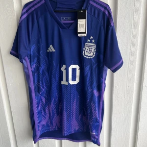 Argentina Messi VM 2022 fotbollströja - Säljer en officiell Argentina fotbollströja från VM 2022 med Messi och nummer 10 på ryggen. Tröjan är blå med lila detaljer, har korta ärmar och ett snyggt grafiskt mönster framtill. Adidas-logga och AFA-emblem på bröstet, tillverkad i lätt polyester.