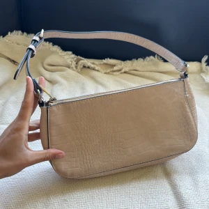 Beige axelväska i croco-imitation - Snygg beige axelväska med croco-mönstrad yta och justerbar rem. Väskan har en stilren, avlång form och stängs med dragkedja upptill. Perfekt storlek för det viktigaste och ger en cool vibe till din outfit. Från NA-KD.