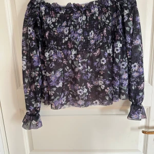 Blommig offshoulder topp Gina Tricot - Supersöt offshoulder blus från Gina Tricot med blommigt mönster i lila, svart och rosa toner. Topp i transparent polyester med smock upptill och volangdetaljer vid ärmslut. Perfekt för dig som gillar romantisk och trendig stil.