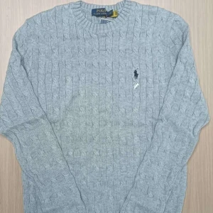 Grå stickad tröja från Polo Ralph Lauren - Säljer en grå stickad tröja från Polo Ralph Lauren med klassiskt kabelstickat mönster och rund hals. Tröjan har långärmade ribbade muddar och den ikoniska Polo-loggan broderad på bröstet. Perfekt för dig som gillar stilren och tidlös design. Icke använd, Säljer för det inte är min still längre