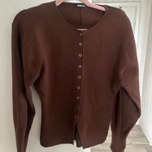 Brun kofta från Gina Tricot - Snygg brun kofta från Gina Tricot i storlek S. Koftan har rund halsringning, långa ärmar och knappar hela vägen framtill. Materialet är mjukt och stickat, perfekt för lager-på-lager under kyliga dagar. Enkel och stilren design som funkar till många outfits.