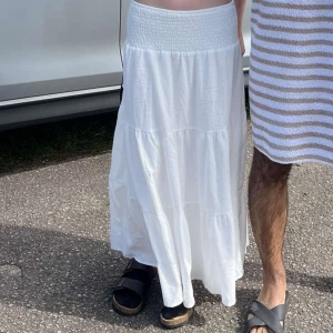 Vit långkjol med volang - Säljer en vit långkjol med bred smock i midjan och flera volanger nedtill. Kjolen är luftig och har en mjuk, flowy känsla som passar perfekt till varma dagar. Enkel och stilren design som är lätt att matcha med olika toppar.