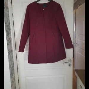 Vinröd kappa från vero moda, storlek 40 - Snygg vinröd kappa från Jyothi i storlek 40. Jackan har en stilren design med raka linjer, diskreta fickor och långärmade ärmar. Materialet är tjockt och känns som ull, med ett slätt foder på insidan. Perfekt för dig som gillar klassisk och enkel stil. Obs: något nopprig