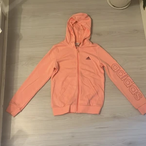 Adidas rosa hoodie med dragkedja - Snygg ljusrosa hoodie från Adidas med dragkedja framtill och huva. Stor Adidas-logga tryckt på ena ärmen och mindre logga på bröstet. Jackan har två fickor och är tillverkad i mjuk bomullsmix som är skön att ha på sig. Perfekt för chill eller träning.