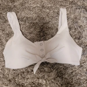 Vit bikiniöverdel från Censored, S - Supersnygg vit bikiniöverdel från New Yorker i storlek S. Modellen har breda axelband, ribbad struktur och en cool knytning framtill med dekorativa knappar.