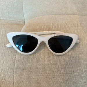 Vita cat eye solglasögon från Mango - Snygga vita solglasögon från Mango med trendig cat eye-form och svarta glas. Ramen är i blank plast och ger en cool retrovibe. Perfekt accessoar för att lyfta din stil och sticka ut i mängden.