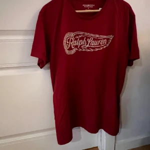 Röd Ralph Lauren t-shirt Denim & Supply - Snygg röd t-shirt från Ralph Lauren Denim & Supply med stort tryck framtill i vitt. Klassisk rund hals och korta ärmar. Tillverkad i 100% bomull för skön känsla. Perfekt för dig som gillar streetwear och vill ha en cool logga på bröstet. Passar L.