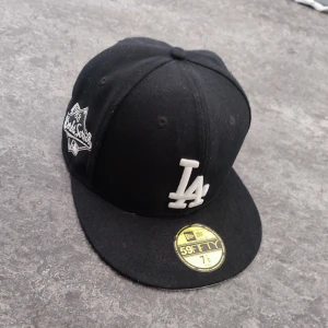 Svart La keps - Snygg svart keps från New Era med LA-logga i vitt framtill. Kepsen är en 59FIFTY-modell med en platt skärm och broderad detalj på sidan. Perfekt för en sportig look.