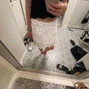 Vit blommig kjol - Supersöt vit omlottkjol med små röda blommor och volangkant. Säljer då den är för liten och det är därför jag inte dragit upp hela dragkedjan där bak🥰💓