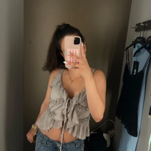 Grå omlott volangtop med snörning - Säljer en grå omlott topp med volanger och snörning framtill. Toppen är croppad och har v-ringning samt smala axelband. Materialet är lätt och känns som bomull eller syntet, perfekt för varma dagar. Snygg att matcha med jeans eller kjol.