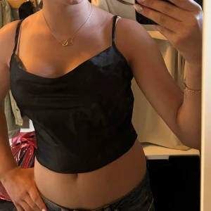 Svart croppad linnetopp från SHEIN - Säljer en svart croppad linnetopp från SHEIN i storlek S. Toppen har tunna justerbara axelband och en snygg draperad urringning framtill. Materialet är glansigt och känns lätt mot huden, perfekt för att lyfta vilken outfit som helst.