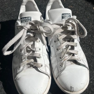 Adidas Stan Smith vita sneakers - Säljer ett par klassiska Adidas Stan Smith sneakers i vitt med mörkblå detalj på hälen. Skorna har snörning, perforerade sidor och platt sula. Materialet är skinn och de har rund tå. Dessa är väl använda! Dom har gått sönder lite i skinnet på skorna lite överallt men jag tycker fortfarande att dom är superfina. Dom är i storlek 35,5. (För att göra det väldigt tydligt så dessa skor har gått sönder i skinnet lite överallt!) Kan skicka noggrannare bilder vid intresse! PRISET ÄR INTE FAST!💙💙💙
