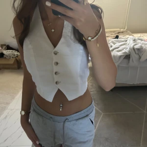 Vit croppad topp med knyt i nacken - Säljer en vit croppad topp med snygg v-ringning och silvriga knappar framtill. Västen har öppen rygg och knytband i nacken för en cool och trendig look. Perfekt att styla till jeans eller kjol för en fräsch vibe. Köpte den för 399 hos ginatricot💕