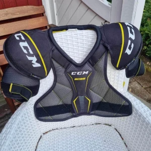 CCM 3092 skyddsväst svart/gul - Säljer en sportig skyddsväst från CCM, modell 3092. Västen är svart med gula och grå detaljer, har breda axelskydd och justerbara remmar. Materialet är syntet med meshpaneler för ventilation. Perfekt för hockey eller liknande sporter. Bra skick, använt 1-2 sesonger. CCM JR LARGE. Cirka 400kr nypris.