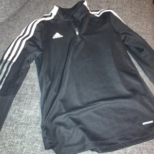 Svart Adidas half zip tröja S - Svart långärmad tröja från Adidas med vita ränder längs ärmarna och Adidas-logga på bröstet. Tröjan har half zip framtill och är gjord i Primegreen polyester, perfekt för träning eller chill. Sportig och snygg passform.