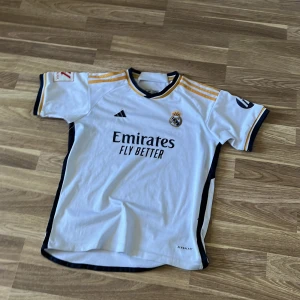 Real Madrid fotbollströja Bellingham 5 - Säljer en vit Real Madrid matchtröja med Bellingham 5 på ryggen. Tröjan har gula och svarta detaljer, korta ärmar och La Liga-märke på ärmen. Adidas-logga och klubbmärke framtill, samt sponsortryck. Passar till nästan allt! Nypris=799kr Mitt pris=149kr! Vid fundering kring prudukten eller annat så är det bara att dma oss😊.          M.V.H//////// Avn Closet🍾🍾