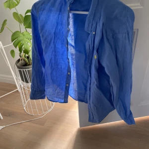 Blå linneskjorta från Polo Ralph Lauren - Säljer en klassisk blå skjorta i linne från Polo Ralph Lauren. Skjortan har lång ärm, knappar framtill och den ikoniska broderade loggan på bröstet. Perfekt för en stilren och avslappnad look.