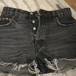 Svarta jeansshorts med fransar  - Snygga svarta jeansshorts från Zara med hög midja och råa fransade kanter. Klassisk femficksmodell med knappar framtill och slitna detaljer för en cool look.
