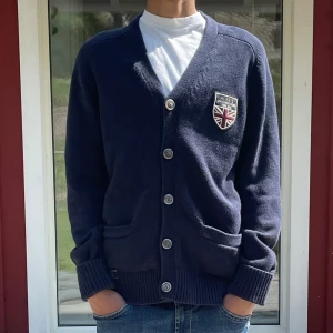 Mörkblå Morris kofta - Morris mörkblå cardigan kofta med v-ringning och knappar framtill. På vänster bröst sitter ett broderat emblem. Pris kan diskuteras.