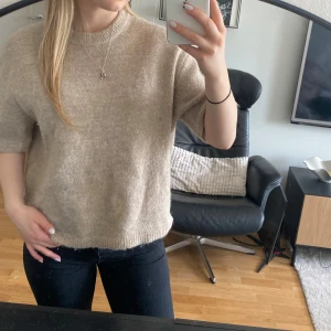 Beige stickad tröja med kort ärm - Säljer en beige stickad tröja med korta ärmar och rund hals. Tröjan har en avslappnad passform och är perfekt för lager-på-lager. Supermjuk och enkel att matcha med olika outfits.