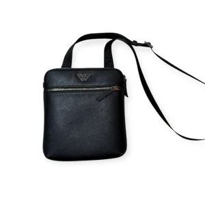 Svart axelväska från Emporio Armani - Snygg svart axelväska från Emporio Armani med justerbar axelrem och silverfärgad dragkedja framtill. Väskan har diskret mönstrad yta och logotyp i metall på framsidan. Perfekt för att bära det viktigaste med stil.