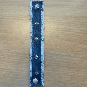 Jeansarmband med nitar - Coolt armband i blått jeansmaterial med fransiga kanter och silverfärgade nitar. Stängs med tryckknappar i båda ändar. Perfekt accessoar för en edgy stil.