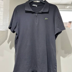 Mörkgrå pikétröja med dragkedja från Lacoste - Säljer en stilren mörkgrå pikétröja från Lacoste med klassisk krage och dragkedja framtill. Tröjan har korta ärmar och den ikoniska krokodilloggan på bröstet. Perfekt för en avslappnad men snygg look.