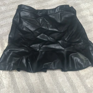 Svart volangkjol i skinnimitation från Kappahl - Säljer en svart kort kjol i skinnimitation från Kappahl. Kjolen har en volang nertill och dragkedja i sidan för enkel påklädning. Perfekt för dig som gillar trendiga och coola plagg med lite edge. Använd ganska många gånger.