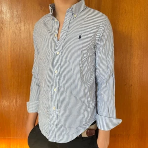 Blåvit randig skjorta från Ralph Lauren - Säljer en klassisk blåvit randig skjorta från Ralph Lauren med broderad logga på bröstet. Skjortan har lång ärm, knappar framtill och button-down krage. Perfekt för en stilren look. Den sitter som storlek xs men är i storlek 16 (modellen är 173cm) skriv för annan info/bilder. 