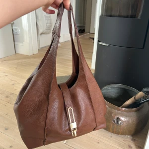 Brun handväska i skinn med gulddetalj från Ullared  - Säljer en brun handväska i äkta skinn med snygg guldfärgad spänndetalj framtill. Väskan har dubbla handtag och en mjuk, rymlig form som gör den perfekt för vardagens alla prylar. Stilren och klassisk design som passar till många outfits.