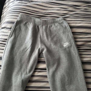 Grå mjukisbyxor från Nike - Grå mjukisbyxor från Nike med resår i midjan och diskret Nike-logga på vänster ben. Byxorna har en bakficka och är tillverkade i mjukt material som passar perfekt för avslappnade dagar eller träning.