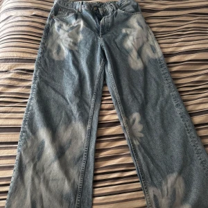 Blommiga vida jeans från Zara - Säljer ett par unika blå jeans från Zara med stora blommiga mönster i vitt. Byxorna har en vid passform och klassisk femficksdesign. Perfekta för dig som vill sticka ut med en cool och trendig look.