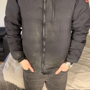 Svart dunjacka från Canada Goose - Säljer en svart dunjacka från Canada Goose med klassisk logga på ärmen. Jackan har dragkedja framtill och lång ärm. Perfekt för kyliga dagar och har en stilren, enkel design. På armen är jackan lite nopprig men inget man tänker på när jackan sitter på. Jackan är lite små skitig på bilderna, men är tvättad så är som ny igen. Jackan är ifrån johnells.