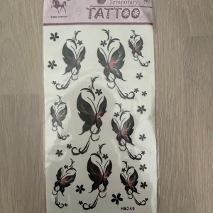 Svarta och rosa fjärilsmotiv temporära tatueringar - Ett ark med temporära tatueringar i form av svarta fjärilar med rosa detaljer och små blommor. Perfekt för att dekorera huden tillfälligt med snygga motiv. Lätta att applicera med vatten och håller några dagar.