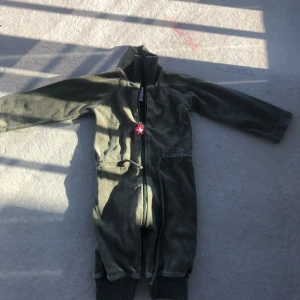 Olivgrön mjukisdress med dragkedja - Olivgrön mjukisdress i onepiece-stil med hel dragkedja framtill och ribbade muddar vid benslut. Dressen har snörning i midjan och en röd dragkedjedekor med vit stjärna. Perfekta för chill eller lek.