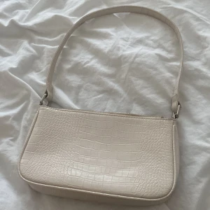 Beige axelväska med krokodilpräglat mönster - Säljer en stilren beige axelväska med krokodilpräglat mönster. Väskan har en enkel design med dragkedja upptill och ett smidigt axelband. Perfekt för att lyfta vilken outfit som helst.
